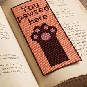 K&ouml;nnte beinhalten: Handgemachter Lesezeichen mit dem Text "You pawsed here" und einem Pfotenabdruck-Design. Das Lesezeichen ist pfirsichfarben mit einem dunkelbraunen Rand und Pfotenabdruck, auf den Seiten eines offenen Buches.