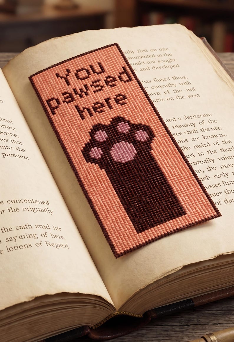 K&ouml;nnte beinhalten: Handgemachter Lesezeichen mit dem Text "You pawsed here" und einem Pfotenabdruck-Design. Das Lesezeichen ist pfirsichfarben mit einem dunkelbraunen Rand und Pfotenabdruck, auf den Seiten eines offenen Buches.