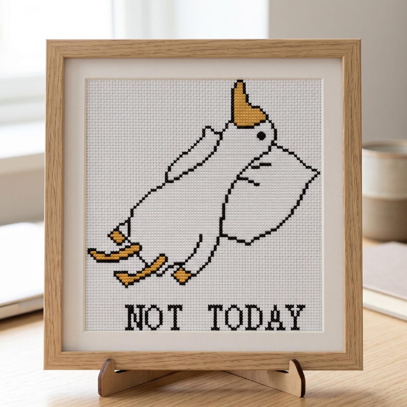 May include: Cross-stitch artwork in a light wood frame. The design features a white cartoon goose with yellow accents, lying on a pillow. The text "NOT TODAY" is stitched below the goose. The artwork is displayed on a wooden stand.