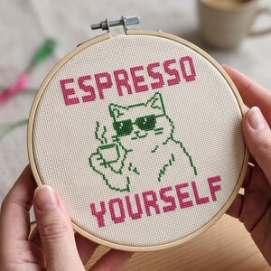 Puede incluir: Un bastidor de bordado circular de madera con un dise&ntilde;o de punto de cruz sobre tela color crema. El dise&ntilde;o muestra un gato con contorno verde y gafas de sol verde oscuro, sosteniendo una taza, con el texto rosa ESPRESSO y YOURSELF.