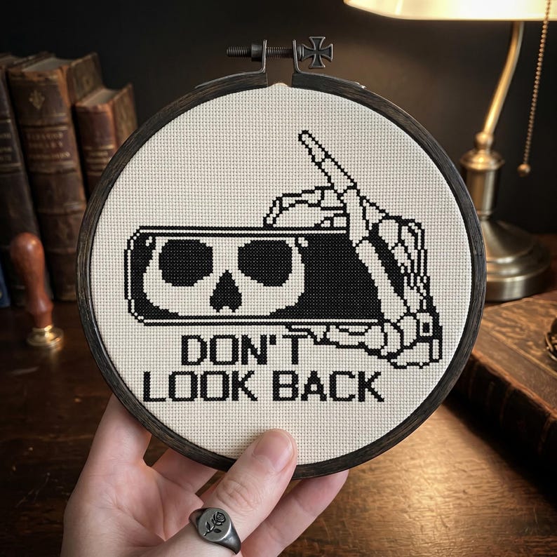 May include: A circular embroidery hoop containing a cross-stitch design on off-white fabric. The design features a black and white skeleton hand holding a smartphone displaying a skull icon, with the text DON'T LOOK BACK printed below.