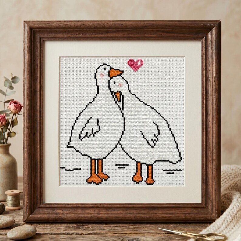 Puede incluir: Obra de arte de punto de cruz en un marco de madera. La imagen presenta dos gansos blancos con contornos negros, picos y patas naranjas, y un coraz&oacute;n rosa encima. La obra se exhibe sobre una superficie de madera.