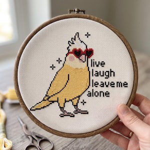 K&ouml;nnte beinhalten: Ein runder Holzstickrahmen mit wei&szlig;em Stoff, der ein Kreuzstichmotiv eines gelben Nymphensittichs mit roten herzf&ouml;rmigen Sonnenbrillen zeigt. Der schwarze Text lautet: live laugh leave me alone. Kleine schwarze Sternsymbole umgeben den Vogel.