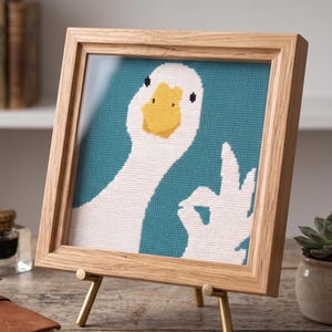 Puede incluir: Una obra de arte en punto de cruz enmarcada que presenta un ganso blanco con pico amarillo y ojos negros, haciendo un gesto de la mano de 'ok'. La obra de arte est&aacute; sobre un fondo verde azulado y enmarcada en un marco de madera marr&oacute;n claro, exhibida en un peque&ntilde;o caballete dorado.