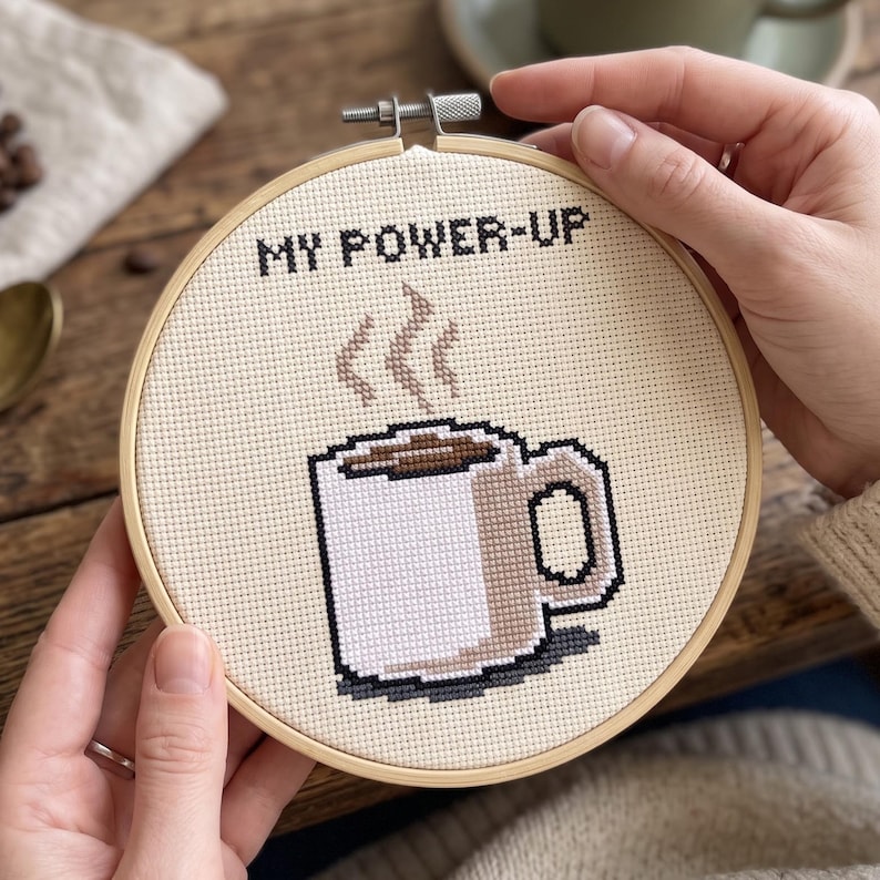 Puede incluir: Un bastidor de bordado circular de madera que contiene un dise&ntilde;o de punto de cruz sobre tela color crema. El dise&ntilde;o presenta una taza de caf&eacute; blanca con l&iacute;quido marr&oacute;n y vapor, junto al texto bordado en negro MY POWER-UP.