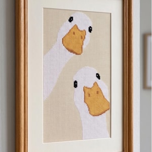 May include: A framed cross-stitch embroidery featuring two white geese with yellow beaks and black eyes against a light beige background. The artwork is enclosed in a light-toned wooden frame with a cream-colored mat board border.