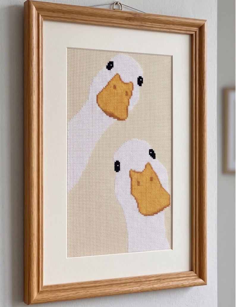 May include: A framed cross-stitch embroidery featuring two white geese with yellow beaks and black eyes against a light beige background. The artwork is enclosed in a light-toned wooden frame with a cream-colored mat board border.