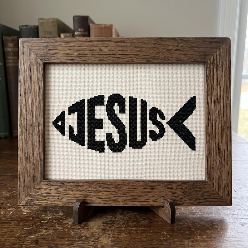 Puede incluir: Un bordado en punto de cruz que presenta la palabra JESUS dispuesta en forma de pez, cosida con hilo negro sobre un fondo de tela color crema. La pieza est&aacute; encerrada en un marco rectangular de madera marr&oacute;n con veta visible.