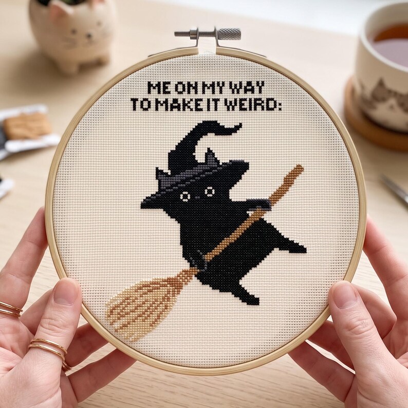 Puede incluir: Un bastidor de bordado circular de madera que contiene un dise&ntilde;o de punto de cruz sobre tela color crema. El dise&ntilde;o muestra un gato negro con sombrero de bruja montado en una escoba marr&oacute;n, con el texto negro ME ON MY WAY TO MAKE IT WEIRD: arriba.