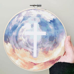 Pode incluir: Bordado em ponto cruz num bastidor de madeira, retratando uma cruz branca sobre um fundo rodopiante de azuis, roxos e tons de p&ecirc;ssego. O design evoca um c&eacute;u nublado, criando um tema espiritual.
