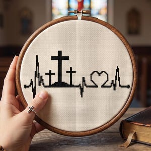 May include: A circular wooden embroidery hoop containing cream-colored Aida fabric with a black cross-stitch design. The pattern features a heartbeat line, three crosses on a small hill, and a heart shape, all rendered in black thread.