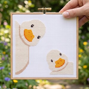K&ouml;nnte beinhalten: Ein quadratisches Kreuzstich-Stickbild mit zwei hellbeigen Enten mit leuchtend orangefarbenen Schn&auml;beln auf wei&szlig;em Stoffhintergrund, befestigt in einem quadratischen hellen Holzrahmen mit einer Metallspannschraube oben in der Mitte.