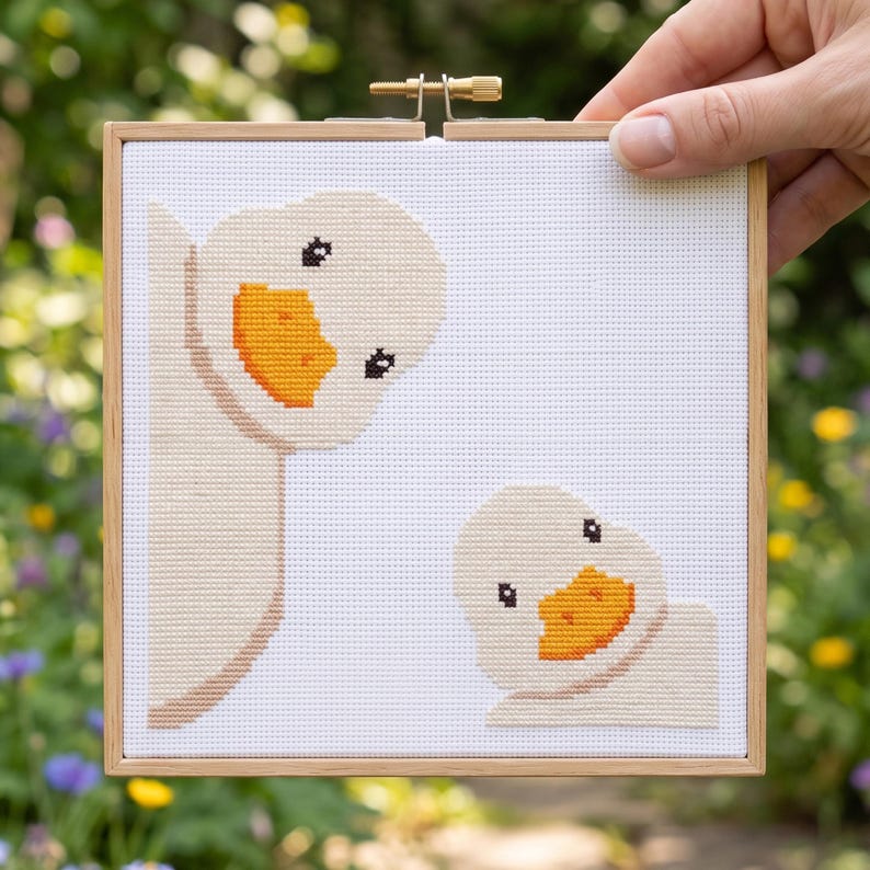 K&ouml;nnte beinhalten: Ein quadratisches Kreuzstich-Stickbild mit zwei hellbeigen Enten mit leuchtend orangefarbenen Schn&auml;beln auf wei&szlig;em Stoffhintergrund, befestigt in einem quadratischen hellen Holzrahmen mit einer Metallspannschraube oben in der Mitte.