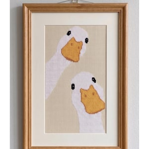 Op de afbeelding: Ingelijst kruissteekpatroon met twee witte ganzen met gele snavels en zwarte ogen. Het kunstwerk staat tegen een beige achtergrond en is ingelijst in een lichtbruine houten lijst. De tekst "CROSS STITCH PATTERN - INSTANT DOWNLOAD" staat bovenaan.