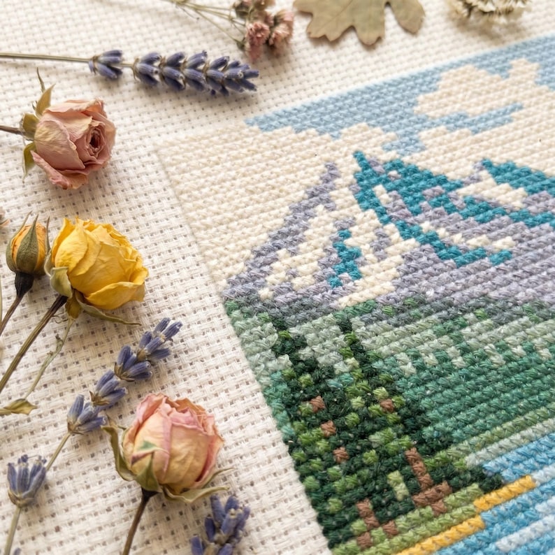 Puede incluir: Un bordado de punto de cruz sobre tela Aida blanca que representa un paisaje de monta&ntilde;a con picos azules, pinos verdes y un cielo azul claro. La pieza est&aacute; decorada con ramas de lavanda seca y capullos de rosa secos rosados y amarillos.