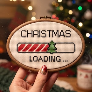 Pu&ograve; includere: Un telaio da ricamo ovale in legno contenente un tessuto bianco con un disegno a punto croce. Il testo recita CHRISTMAS LOADING... con una barra di caricamento a strisce diagonali rosse e bianche e un'icona di pino verde al centro.