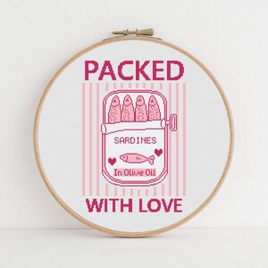 Pu&ograve; includere: Un telaio da ricamo a punto croce con un design rosa e bianco. Il design include le parole "PACKED WITH LOVE" e un'illustrazione di una scatoletta di sardine con le parole "SARDINES In Olive Oil".