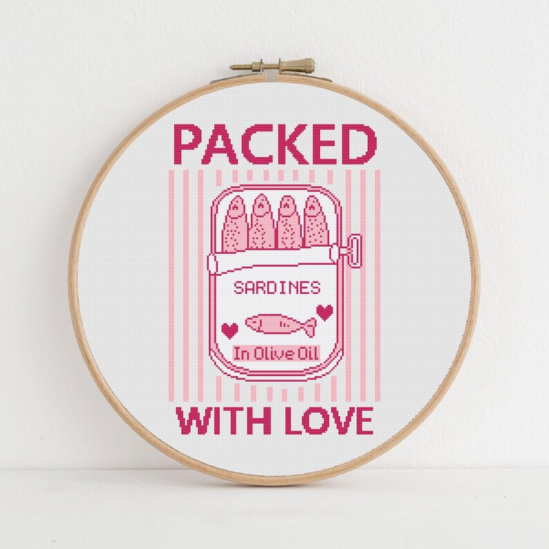Pu&ograve; includere: Un telaio da ricamo a punto croce con un design rosa e bianco. Il design include le parole "PACKED WITH LOVE" e un'illustrazione di una scatoletta di sardine con le parole "SARDINES In Olive Oil".