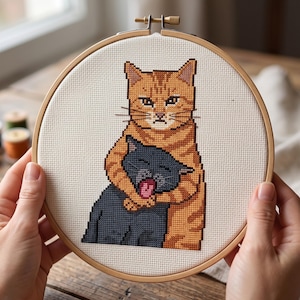 Puede incluir: Un proyecto de bordado en punto de cruz dentro de un bastidor de madera circular que muestra a un gato atigrado naranja abrazando a un gato gris oscuro m&aacute;s peque&ntilde;o. El gato naranja tiene una expresi&oacute;n seria y el gris tiene la boca abierta.