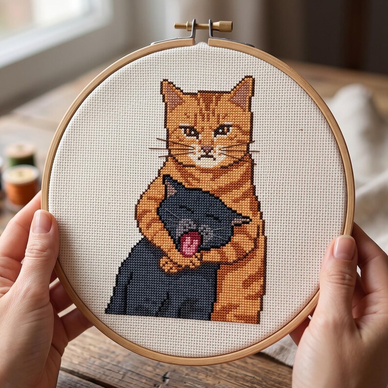 Puede incluir: Un proyecto de bordado en punto de cruz dentro de un bastidor de madera circular que muestra a un gato atigrado naranja abrazando a un gato gris oscuro m&aacute;s peque&ntilde;o. El gato naranja tiene una expresi&oacute;n seria y el gris tiene la boca abierta.