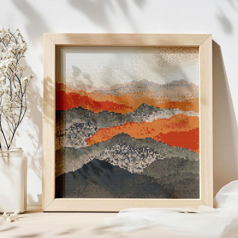 May include: A framed cross-stitch landscape depicting a mountain range in shades of orange, grey, and black. The artwork is presented in a light-coloured wooden frame. The sky is a gradient of white and light grey.