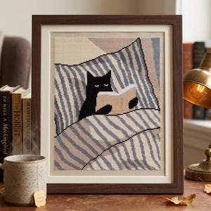 Peut inclure: Une broderie au point de croix encadrée représentant un chat noir allongé dans un lit sous une couverture à rayures bleues et blanches, en train de lire un livre ouvert. L'œuvre est placée dans un cadre en bois brun foncé avec un passe-partout crème.