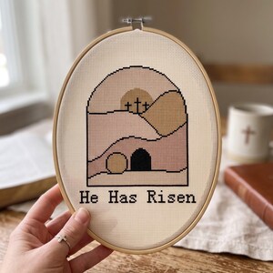 K&ouml;nnte beinhalten: Ein ovaler Stickrahmen mit einem Kreuzstichmotiv auf cremefarbenem Stoff. Das Design zeigt eine Landschaft mit drei Kreuzen auf einem H&uuml;gel, einem Grabeingang und dem Text "He Has Risen" in schwarzem Garn. Die Farbpalette umfasst Beige, Rosa und Braun.