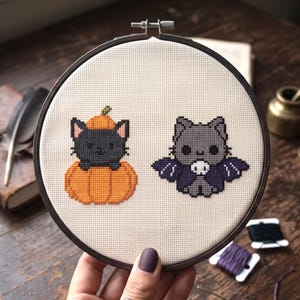 May include: A circular wooden embroidery hoop containing a piece of cream-colored Aida fabric. The fabric features a cross-stitch design of a black cat sitting inside an orange pumpkin and a gray cat wearing a purple bat costume with a skull icon.