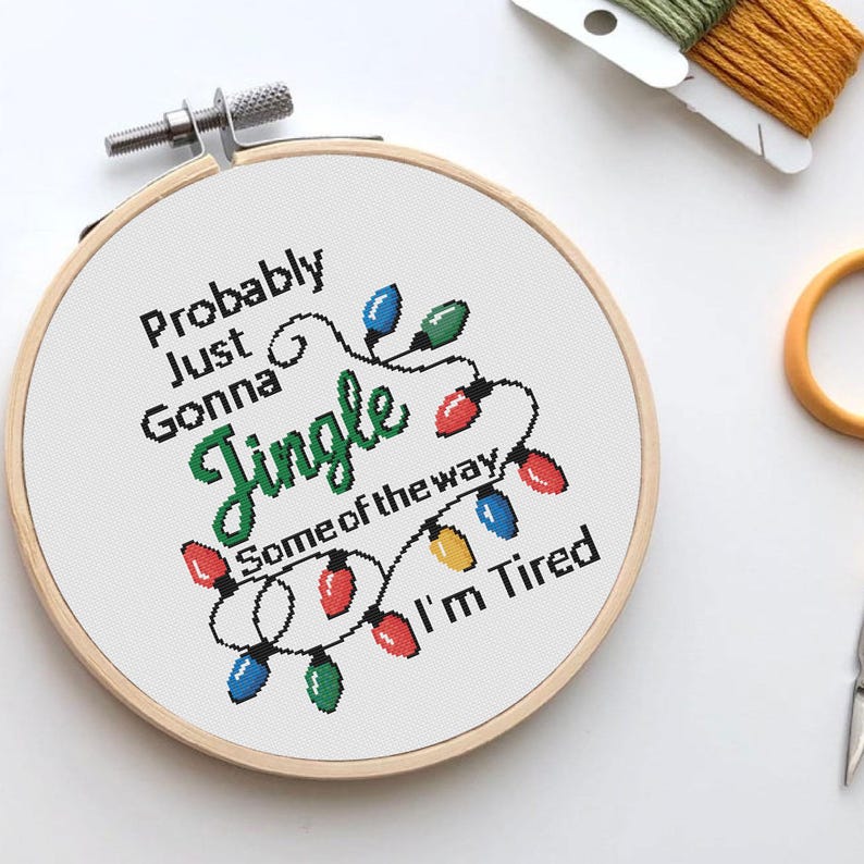 Może przedstawiać: Obręcz do haftu krzyżykowego z napisem "Probably Just Gonna Jingle Some of the way I'm Tired" w kolorze czarnym i zielonym. Projekt zawiera kolorowe lampki choinkowe. Drewniana obręcz jest zabezpieczona metalową klamrą.