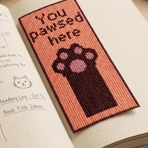 Op de afbeelding: Handgemaakte bladwijzer met de tekst "You pawsed here" en een pootafdruk. De bladwijzer is perzik van kleur met een donkerbruine rand en pootafdruk, geplaatst in een open boek.