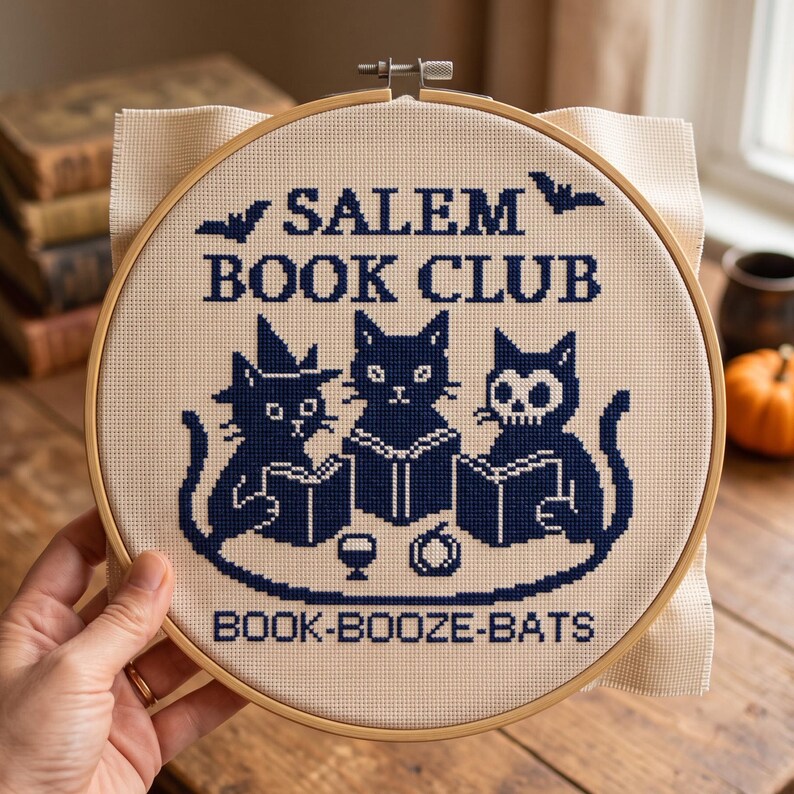 Puede incluir: Un bastidor de bordado circular de madera que contiene un dise&ntilde;o de punto de cruz sobre tela beige. El dise&ntilde;o muestra tres gatos negros leyendo libros, con el texto SALEM BOOK CLUB y BOOK-BOOZE-BATS en hilo azul oscuro, con murci&eacute;lagos.