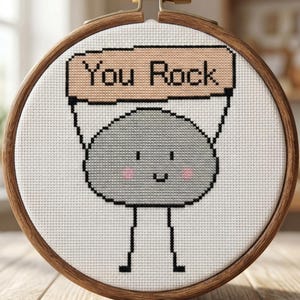 Op de afbeelding: Kruissteekpatroon in een houten borduurring. Het ontwerp toont een grijze rots met een glimlach die een bord vasthoudt met de tekst "You Rock". De tekst "CROSS STITCH PATTERN - INSTANT DOWNLOAD" staat bovenaan.