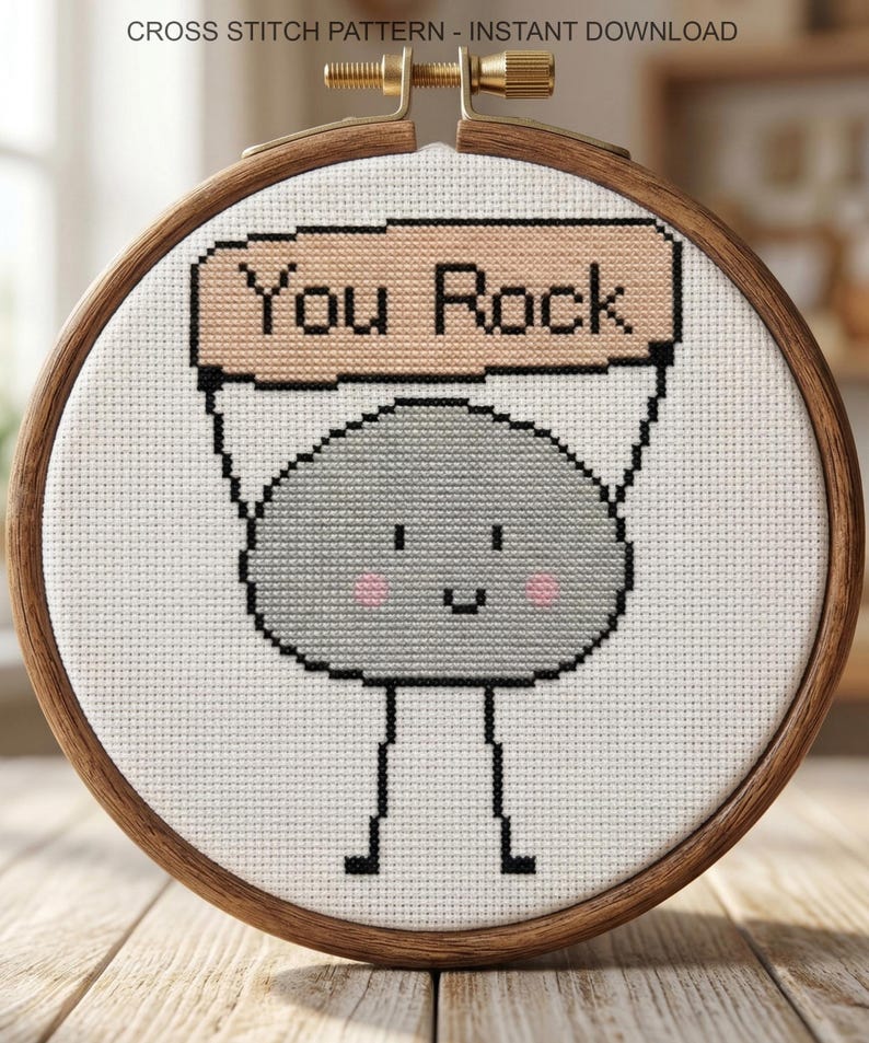 K&ouml;nnte beinhalten: Ein Kreuzstichmuster in einem h&ouml;lzernen Stickrahmen, das einen grauen Felsencharakter mit l&auml;chelndem Gesicht und rosa Wangen zeigt, der ein beiges Schild mit der Aufschrift You Rock h&auml;lt. Der Text steht oben im Bild.