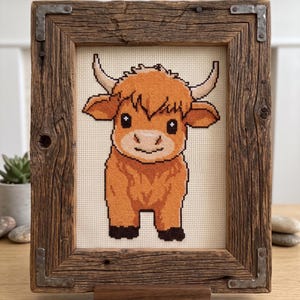May include: Cross-stitch artwork of a Highland cow in a rustic wooden frame. The cow is rendered in shades of orange, brown, and black, with a cream-colored background. The frame is made of reclaimed wood with metal corner brackets and a wooden stand.