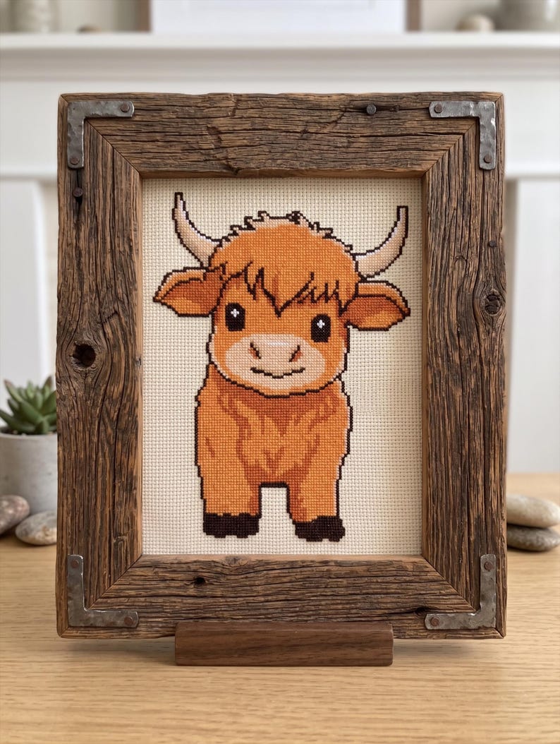 May include: Cross-stitch artwork of a Highland cow in a rustic wooden frame. The cow is rendered in shades of orange, brown, and black, with a cream-colored background. The frame is made of reclaimed wood with metal corner brackets and a wooden stand.