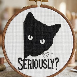 K&ouml;nnte beinhalten: Kreuzstich-Stickerei in einem Holzreifen mit einem schwarzen Katzenkopf auf wei&szlig;em Hintergrund. Der Text "SERIOUSLY?" ist darunter gestickt. Der Reifen hat eine Messing-Einstellschraube.