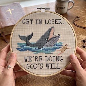 Puede incluir: Un bastidor de bordado circular de madera con un diseño de punto de cruz sobre tela beige. El diseño muestra una ballena gris con la boca abierta en agua azul, una figura pequeña y el texto negro: GET IN LOSER, WE'RE DOING GOD'S WILL.