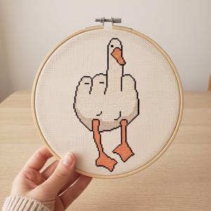 Peut inclure: Un tambour &agrave; broder circulaire en bois contenant un motif au point de croix repr&eacute;sentant une oie blanche au bec et aux pattes orange. L'oie est repr&eacute;sent&eacute;e avec ses ailes lev&eacute;es dans un geste rappelant un majeur, sur un fond en tissu cr&egrave;me.