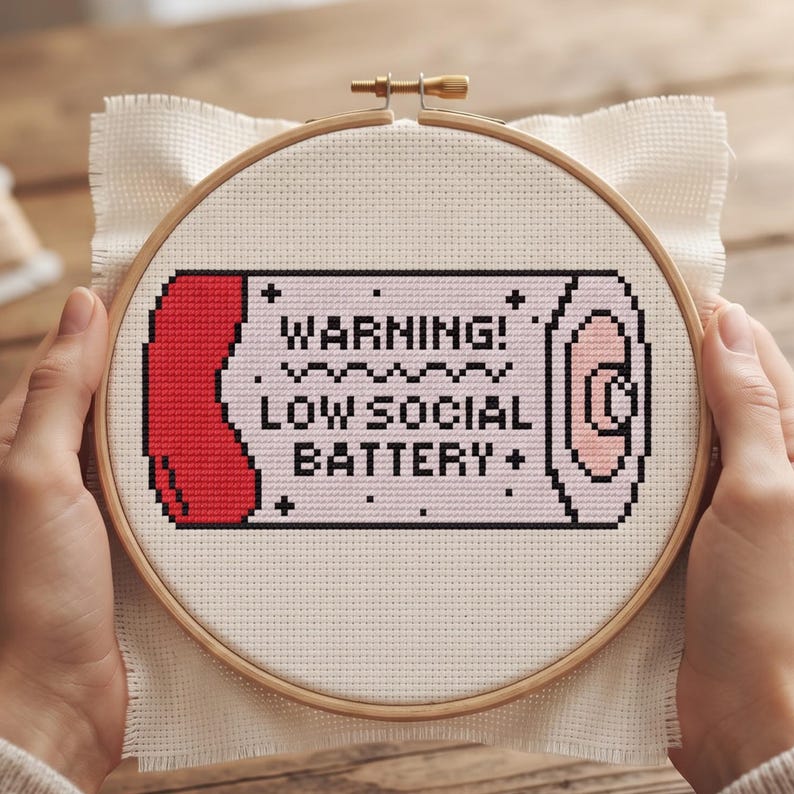 May include: A cross-stitch project in a round wooden embroidery hoop featuring a pixel-art style battery icon. The design includes the black text WARNING! LOW SOCIAL BATTERY on a light pink background with a red end cap and black outlines.