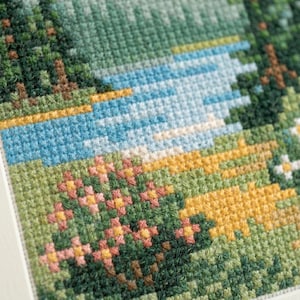 Puede incluir: Un bordado de punto de cruz que representa un paisaje con un r&iacute;o azul que fluye a trav&eacute;s de un bosque de pinos de color verde oscuro. El primer plano presenta una pradera con parches de flores amarillas y rosas, todo en hilo detallado.