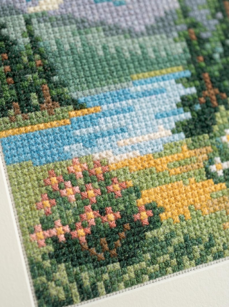 Puede incluir: Un bordado de punto de cruz que representa un paisaje con un r&iacute;o azul que fluye a trav&eacute;s de un bosque de pinos de color verde oscuro. El primer plano presenta una pradera con parches de flores amarillas y rosas, todo en hilo detallado.