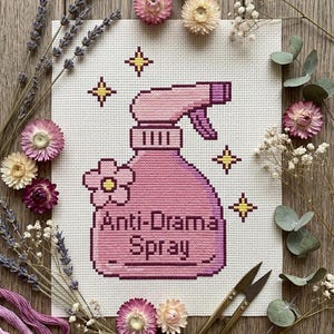Peut inclure: Une broderie au point de croix repr&eacute;sente un flacon pulv&eacute;risateur rose avec l'inscription "Anti-Drama Spray". Le motif est entour&eacute; de fleurs s&eacute;ch&eacute;es, de lavande et de ciseaux dor&eacute;s, cr&eacute;ant une ambiance rustique et artisanale.