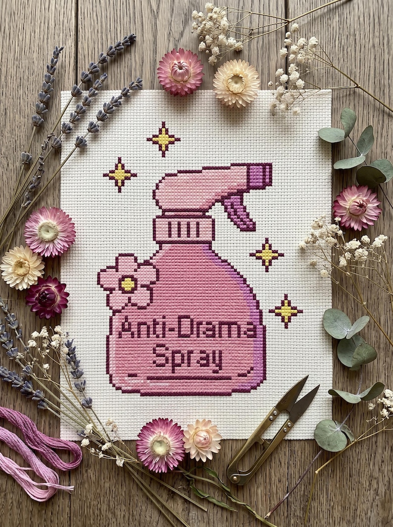 Peut inclure: Une broderie au point de croix repr&eacute;sente un flacon pulv&eacute;risateur rose avec l'inscription "Anti-Drama Spray". Le motif est entour&eacute; de fleurs s&eacute;ch&eacute;es, de lavande et de ciseaux dor&eacute;s, cr&eacute;ant une ambiance rustique et artisanale.