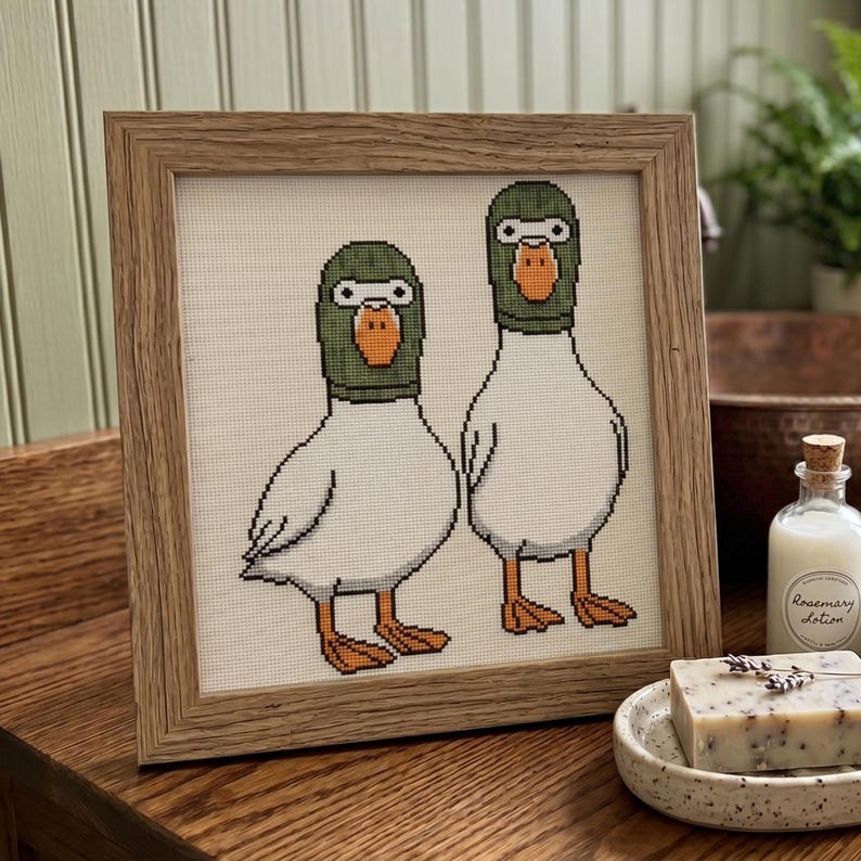 Peut inclure: Un cadre en bois carr&eacute; contenant une broderie au point de croix repr&eacute;sentant deux canards blancs avec des t&ecirc;tes vertes et des becs orange. Les canards sont debout c&ocirc;te &agrave; c&ocirc;te sur un fond en tissu blanc, dans un style pixelis&eacute; aux contours noirs.