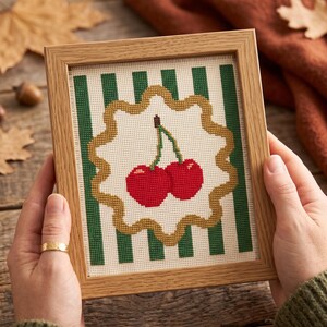 May include: A framed cross-stitch embroidery featuring two bright red cherries with green stems centred on a cream-coloured background. The design is framed by a scalloped gold border and set against a backdrop of vertical dark green stripes.