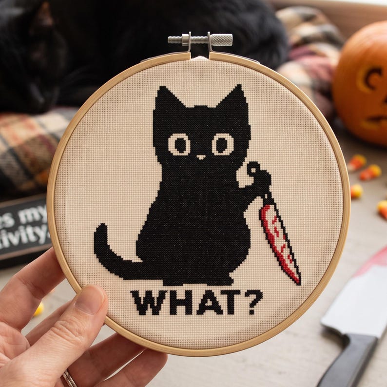 K&ouml;nnte beinhalten: Ein runder Stickrahmen aus Holz mit einem Kreuzstichmotiv auf cremefarbenem Stoff. Das Design zeigt eine schwarze Katze mit gro&szlig;en wei&szlig;en Augen, die ein Messer mit rot gef&auml;rbter Klinge h&auml;lt, darunter der schwarze Schriftzug WHAT?