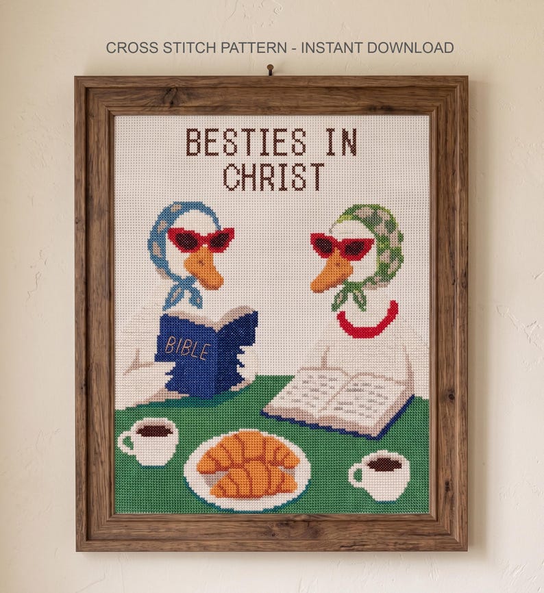 May include: Cross-stitch pattern in a wooden frame featuring two geese wearing sunglasses and headscarves, reading books. The text "BESTIES IN CHRIST" is at the top. A plate of croissants and two cups of coffee are on a green surface.