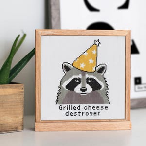 Op de afbeelding: Kruissteekkunst van een wasbeer met een gele feesthoed met witte sterren. De wasbeer heeft een zwart masker en is ingelijst in een houten frame. De tekst "Grilled cheese destroyer" staat onder de wasbeer.