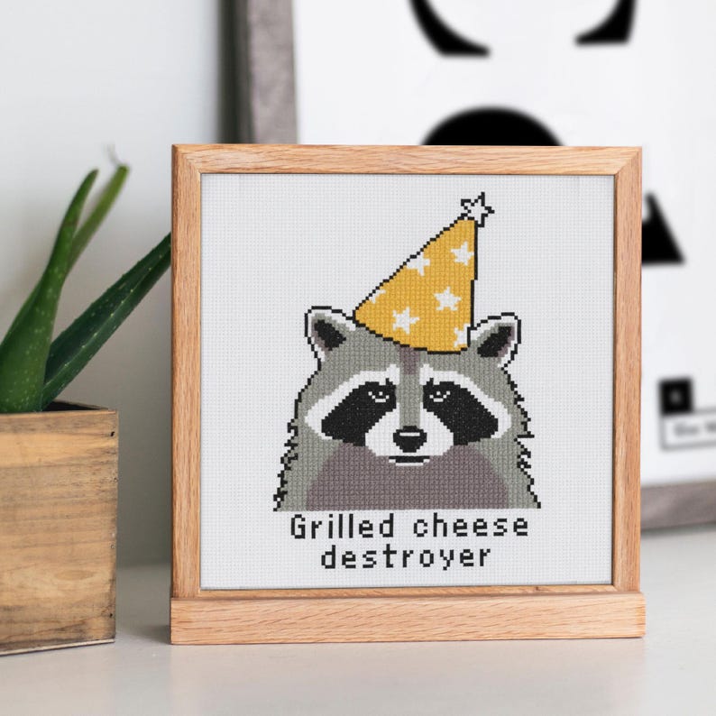 Op de afbeelding: Kruissteekkunst van een wasbeer met een gele feesthoed met witte sterren. De wasbeer heeft een zwart masker en is ingelijst in een houten frame. De tekst "Grilled cheese destroyer" staat onder de wasbeer.