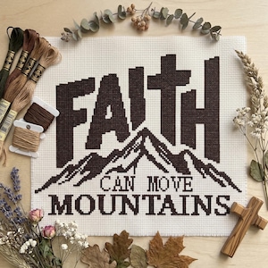 May include: A cross-stitch embroidery on white Aida fabric featuring the dark brown text FAITH CAN MOVE MOUNTAINS with a mountain range graphic. The piece is surrounded by embroidery floss skeins, dried lavender, eucalyptus, and a wooden cross.